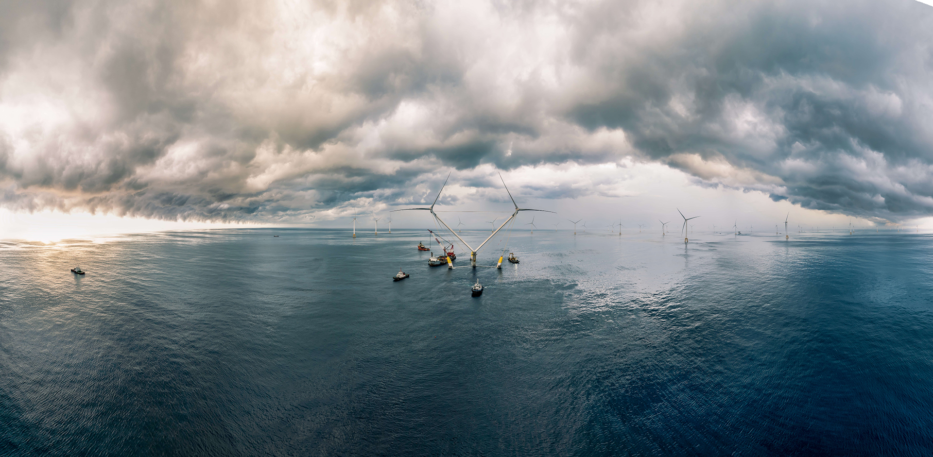 Offshore wind turbines and small vessels on the horizon under a vast, dark sky filled with heavy storm clouds.